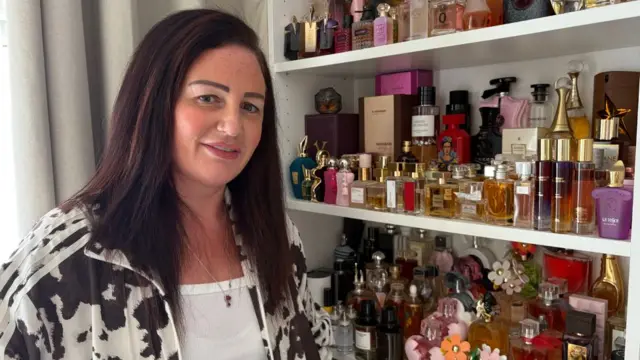 Una mujer con pelo largo oscuro parada frente a un mostrador con perfumes