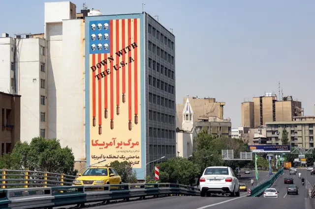 Vehículos transitan por una avenida que tiene a un lado un edificio con un mural que tiene la bandera de EE.UU. y un mensaje que dice