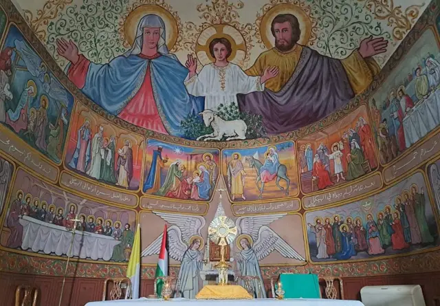 El altar de la parroquia Sagrada Faimilia. No se ve a Cristo crucificado, sino que hay una imagen de Jesús con los brazos abiertos. A un lado tiene a María y al otro a José. 