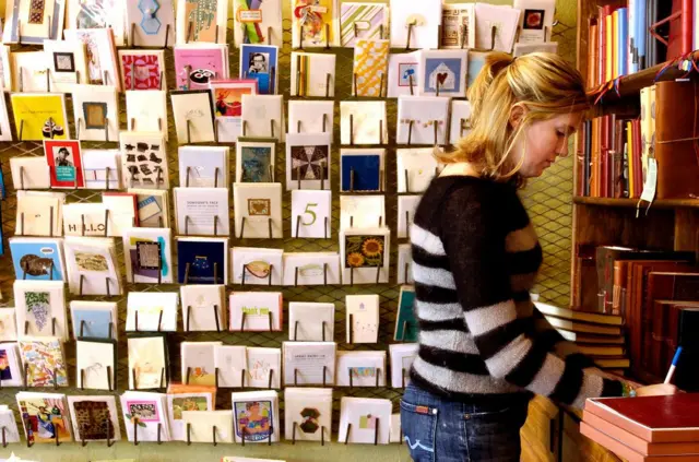Una mujer leyendo una tarjeta frente a los estantes que guardan tarjetas de cumpleaños, bodas, etc.