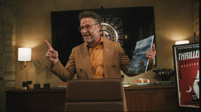 Un hombre habla con los brazos abiertos y un dossier en la mano derecha en lo que parece ser un despacho. A su izquierda se ve un cartel con el texto 