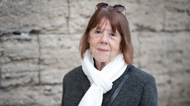 Gisèle Pelicot, una francesa de unos setenta y tantos años, lleva gafas de sol sobre la cabeza, un pañuelo blanco alrededor del cuello y un abrigo oscuro con textura. El fondo presenta un muro de piedra, lo que sugiere un entorno al aire libre o histórico. Gisèle se hizo internacionalmente conocida tras sobrevivir a casi una década de abusos encubiertos por parte de su esposo, Dominique Pelicot. Entre 2011 y 2020, Dominique la drogó e invitó a decenas de hombres a violarla mientras estaba inconsciente, filmando las agresiones.