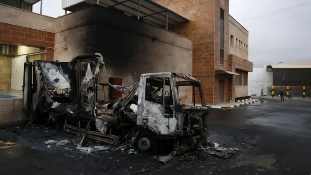 Un las ruinas de camión incendiado frente a un complejo industrial cuyas paredes de ladrillo están manchadas de humo  