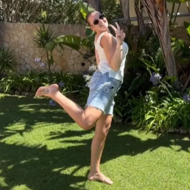 Una mujer con una camiseta blanca sin mangas y uno pantalones cortos posa en un jardín para una foto.