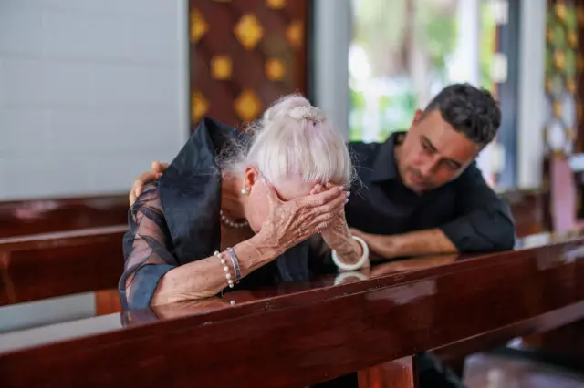 Mujer de pelo canoso con sus manos cubriendo su frente y rostro, está arrodillada en el banco de una iglesia, mientras un hombre más joven la consuela con un brazo sobre su espalda y el otro sobre un brazo.
