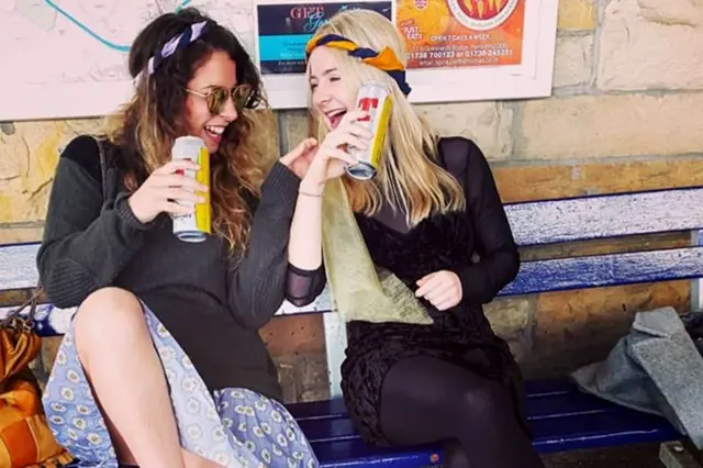 Hazel riendo con una amiga, sentadas en un banco bebiendo cerveza 