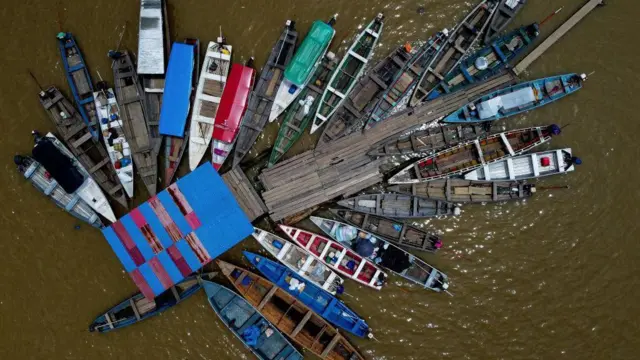 Foto tomada desde arriba que muestra unas 30 pequeñas embarcaciones de madera agrupadas alrededor de un embarcadero de madera, rodeadas por las brillantes aguas marrones del río.