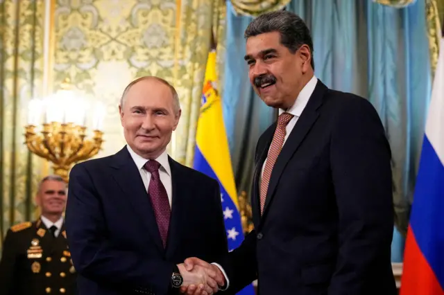 Vladimir Putin (i) y Nicolás Maduro (d) estrechan las manos sonrientes  en Moscú. Ambos lucen traje y corbata oscuros y están rodeados de banderas de Venezuela y Rusia. 