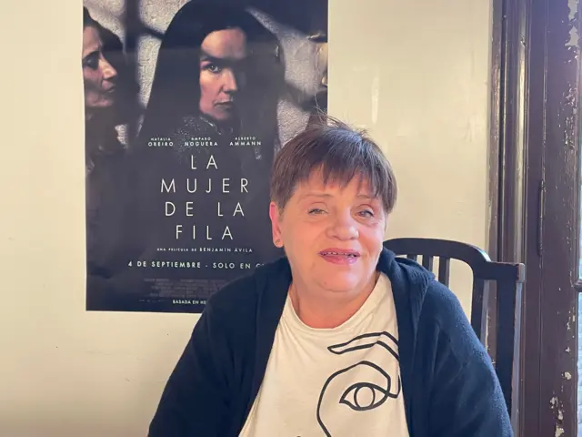 Andrea está sentada y mira a la cámara. Sobre una pared de color claro en el fondo, está pegado un afiche de la película 