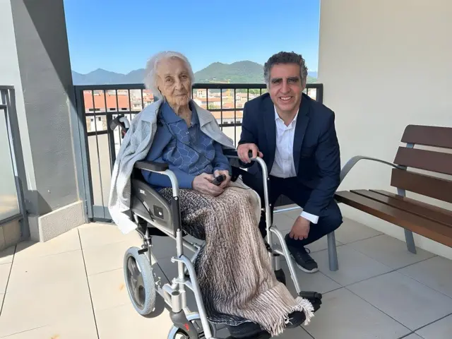 María Branyas sentada en una silla de ruedas, con un saco azul y una manta sobre sus piernas, en un balcón posa junto al doctor Manel Esteller