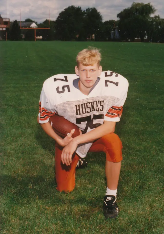 Chris Nowinski, cuando chico, aparece arrodillado en su uniforme de fútbol americano y un balón en la mano, mirando serio a la cámara, en medio de un campo de fútbol americano. 