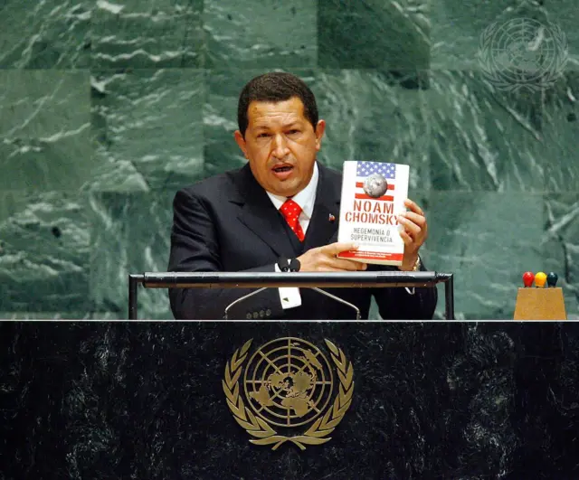 Hugo Chávez viste traje oscuro, camisa blanca y corbata roja. Con sus dos manos puestas hacia la izquierda sostiene un libro blanco de Noam Chomsky. Delante de Chávez, en la parte baja de la imagen se ve la parte frontal del podio de la Asamblea General, de color oscuro.