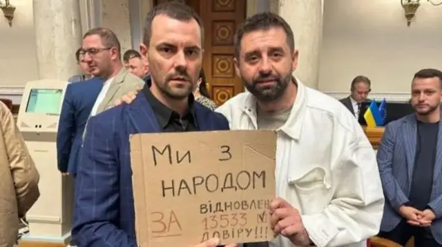 Dos diputados sostienen un cartel de cartón que dice