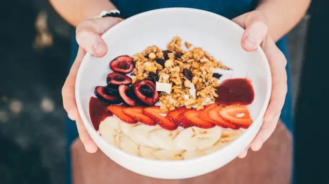 Un primer plano de las manos de una persona sosteniendo un tazón blanco que contiene fresas, banana y muesli.