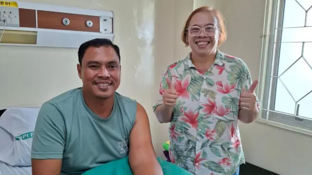 Tri Maharani está de pie a la derecha, con una camisa de flores y gafas de montura rosa, mostrando el pulgar hacia arriba y sonriendo. A su lado hay un paciente, con una camiseta verde, sentado en una cama de hospital.