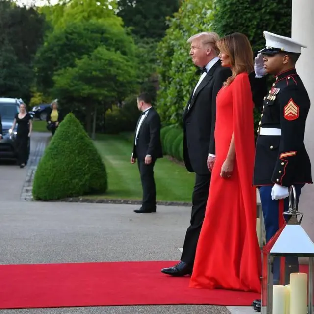Donald y Melania Trump, vestidos de gala, esperan en una alfombra roja a los invitados mientras un soldado saluda junto a ellos.