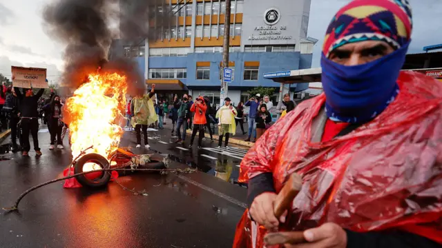 Al fondo, un grupo se reúne alrededor de una fogata de neumáticos en medio de una calle, cerca de un edificio gubernamental, mientras que en primer plano un hombre con un poncho rojo, un pasamontañas azul y un sombrero rojo estampado mira fijamente a la cámara.