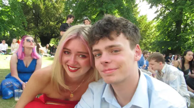 Paloma Shemirani junto a su novio Ander Harris en un parque, rodeados de árboles y otras personas sentadas en el césped