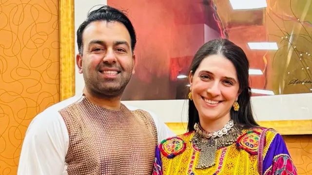 Safi y Sammi están uno al lado del otro y sonríen en su boda. Safi lleva un top blanco con un patrón de celosía marrón en la parte delantera, conocido como Shalwar. Sammi lleva un vestido multicolor. La parte delantera es amarilla con un patrón de flores. Las mangas son moradas, amarillas y rojas.