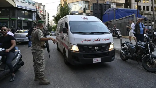 Una ambulancia circula por una calle muy transitada junto a un hombre con uniforme militar y un rifle de asalto.
