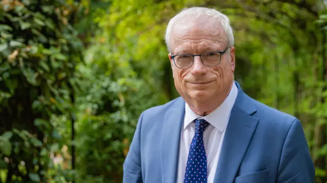 Chris Smith porta un traje azul con una corbata y está parado frente a un jardín de arbustos verdes