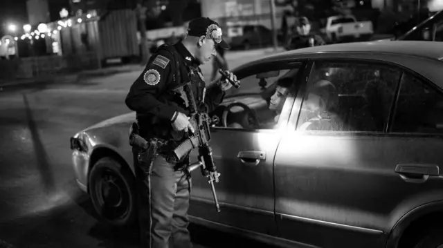 Un agente policial armado alumbra con una linterna a un hombre al volante en un auto detenido. Foto en blanco y negro.