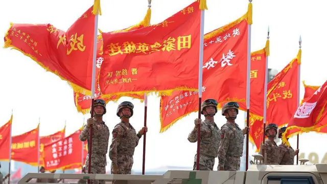 Soldados con uniforme de combate se encuentran en la parte trasera de camiones sosteniendo grandes banderas rojas.