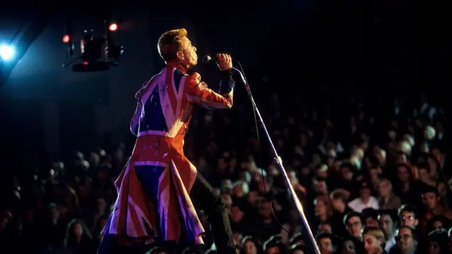 David Bowie actúa frente a un gran público vistiendo un abrigo largo inspirado en la bandera de Reino Unido.