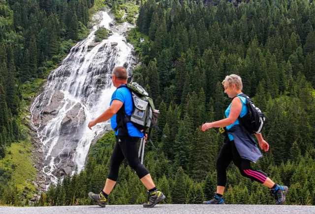 Una pareja de mochileros, con camisas azules y zapatos de montañistas, camina frente a una cascada que cae entre una empinada montaña con árboles verdes