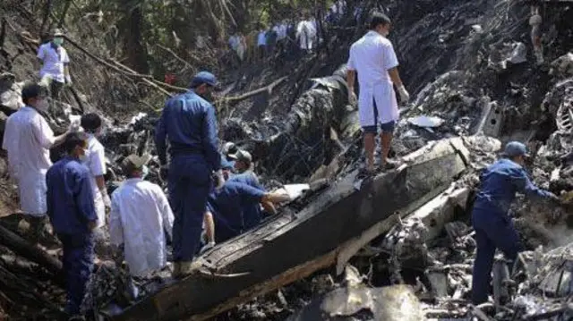 En medio de vegetación, el fuselaje de un avión destruido tras lo que parece ser un accidente aéreo. Alrededor, una decena de hombres vestidos con uniformes observan la escena.