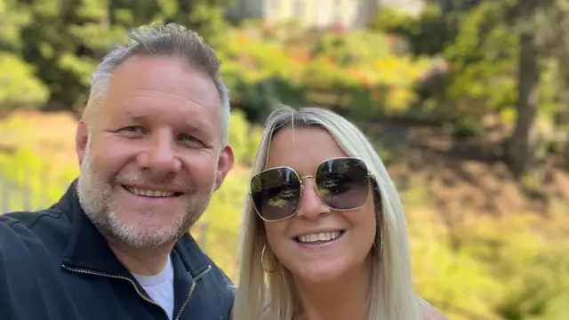Selfie de Ben Giles (izquierda) y su esposa Lindsey (derecha). Ben tiene el pelo corto y canoso, barba gris y ojos azules. Ella sonríe ampliamente a la cámara. Lindsey lleva unas gafas de sol grandes y anchas con montura dorada y pendientes de aro dorados, y sonríe a la cámara. Tiene el pelo largo y rubio brillante. Detrás de ellos se ve césped verde iluminado por el sol, algunos árboles y una casa solariega.