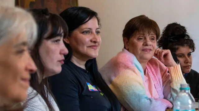 Cinco mujeres de diferentes edades sentadas a lo largo de una mesa mirando hacia una misma dirección. Andrea, la cuarta de izquierda a derecha, luce un saco de colores pastel y se lleva una mano a la cara.