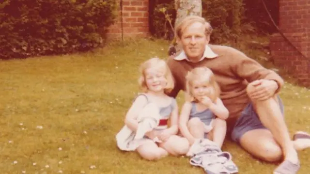Una antigua foto familiar muestra a Smyth con pantalones cortos azules y un suéter marrón, sentado en el césped junto a sus dos hijas pequeñas. Parece un día soleado y todos sonríen a la cámara.