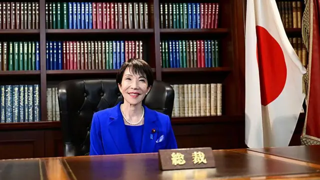 Sanae Takaichi, vestida con un traje azul, sentada en un escritorio de madera sonríe a la cámara. Detrás tiene una biblioteca, a su izquierda la bandera de Japón