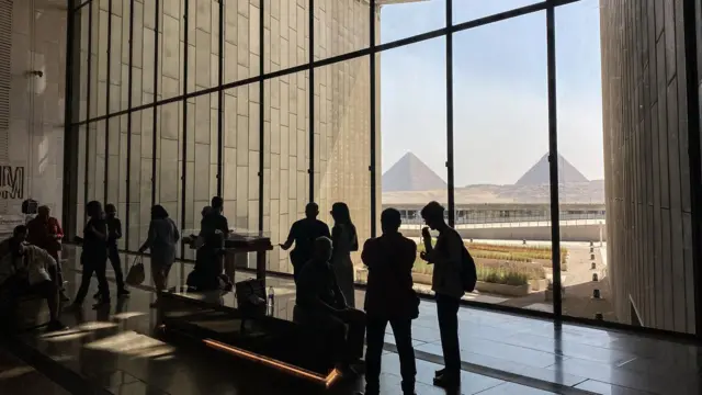 Vista interior del Gran Museo Egipcio, que se alza imponente con las pirámides de Giza de fondo, mientras los visitantes lo recorren.