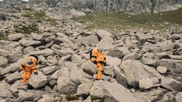 Dos personas vestidas con trajes de astronauta naranjas sobre unas rocas en los Alpes suizos.