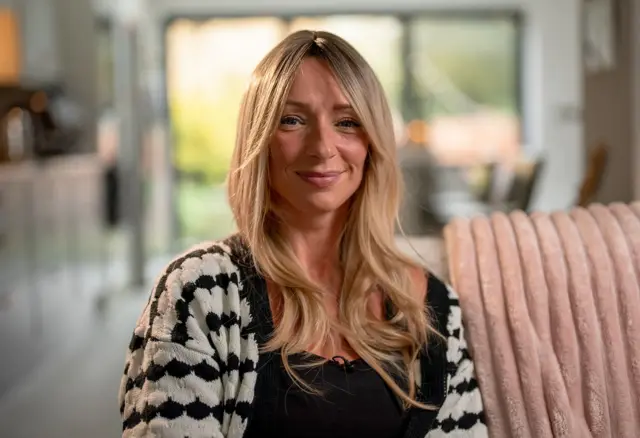 Ellen, una mujer de cabello largo y rubio, está sentada en un sofá sonriendo a la cámara.