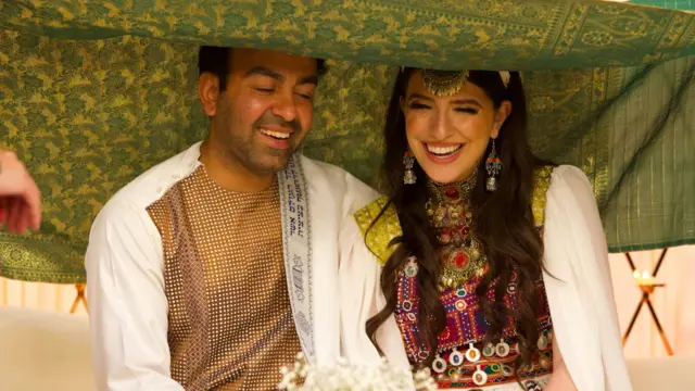 Sammi y Safi sentados bajo un dosel en su boda. Ambos sonríen ampliamente y Sammi tiene los ojos cerrados.
