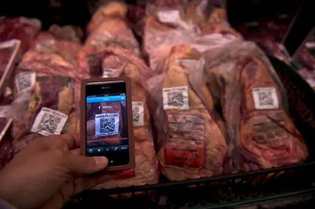 Una bandeja con cortes de carne vacuna envasada que tienen un código QR para obtener su trazabilidad, y delante una persona sostiene con su mano un teléfono y apunta con la cámara a uno de esos códigos.