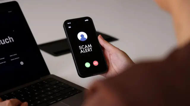 Una persona sostiene un teléfono inteligente que muestra un mensaje de “Alerta de estafa” en la pantalla 