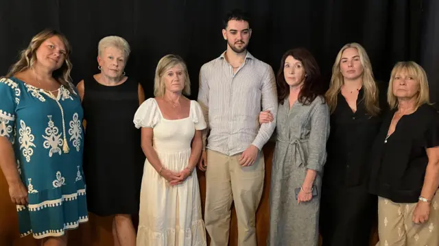 Calum está de pie en medio de la habitación, flanqueado a ambos lados por las familias de Simone, Kirsty y Cheznye.