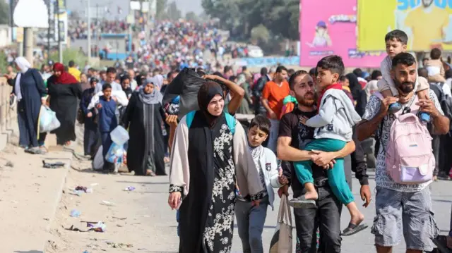 Una multitud de personas camina por una carretera en Gaza el 10 de noviembre de 2023. En primer plano, dos hombres llevan a niños pequeños, uno con un niño a hombros y una mochila rosa atada al pecho. Junto a ellos, una mujer camina junto a un niño mayor. Detrás de ellos, la multitud se extiende por la carretera hasta donde alcanza la vista.