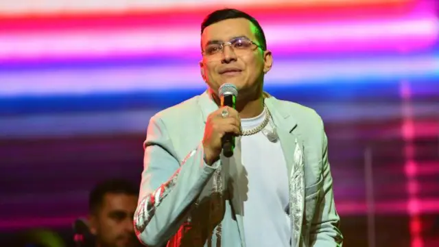 Yeison Jiménez canta sobre un escenario vestido con un atuendo crema y dorado y rodeado de luces de colores.