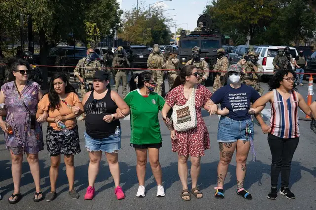 Un grupo de mujeres con ropa de civil y desarmadas se enganchan unas con otras de brazo en una línea frente a agentes migratorios armados con los rostros tapados. 