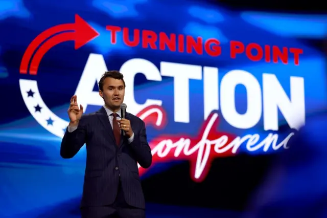 Charlie Kirk en un escenário hablando por un micrófono y con una pantalla detrás en la que se ve el logo de su organización, turning Point.