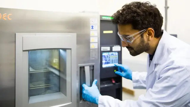 Un investigador con bata blanca, guantes de látex y gafas protectoras opera una especie de horno en el que se encuentran paneles solares.