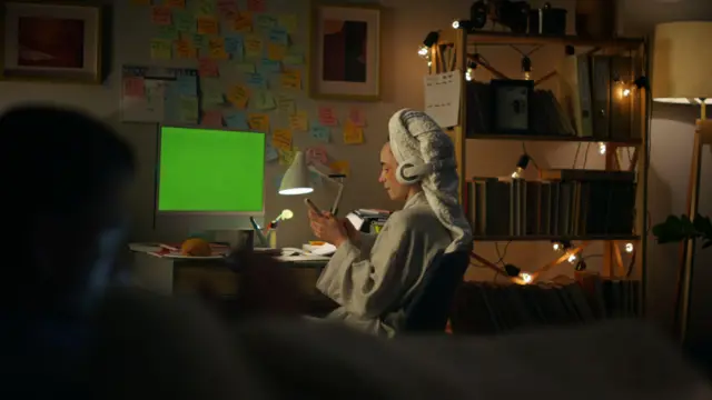 Mujer que se bañó por la noche, en una habitación iluminada solo por unas luces pequeñas, mirando el celular con una toalla en la cabeza y una computadora de fondo