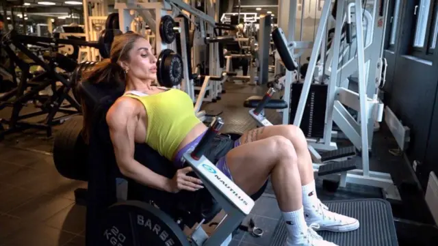 Una mujer en traje deportivo, dentro de un gimnasio, sentada en un aparato para desarrollar los muslos