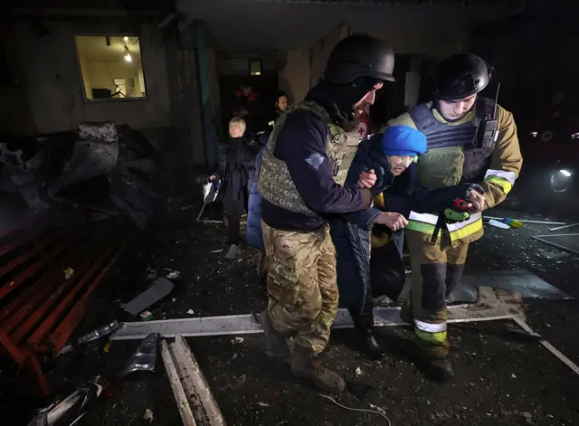 Dos rescatistas ucranianos ayudan a una anciana a ser evacuada del lugar donde un ataque ruso impactó un edificio residencial de gran altura en Járkov, al noreste de Ucrania, el 19 de noviembre de 2025. La mujer luce angustiada y se inclina hacia adelante. Lleva un gorro azul y una chaqueta de invierno.