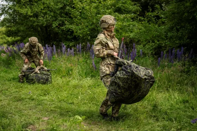 Mujer recluta del ejército danés, vestida con uniforme militar y casco, lleva un rifle y una bolsa grande.